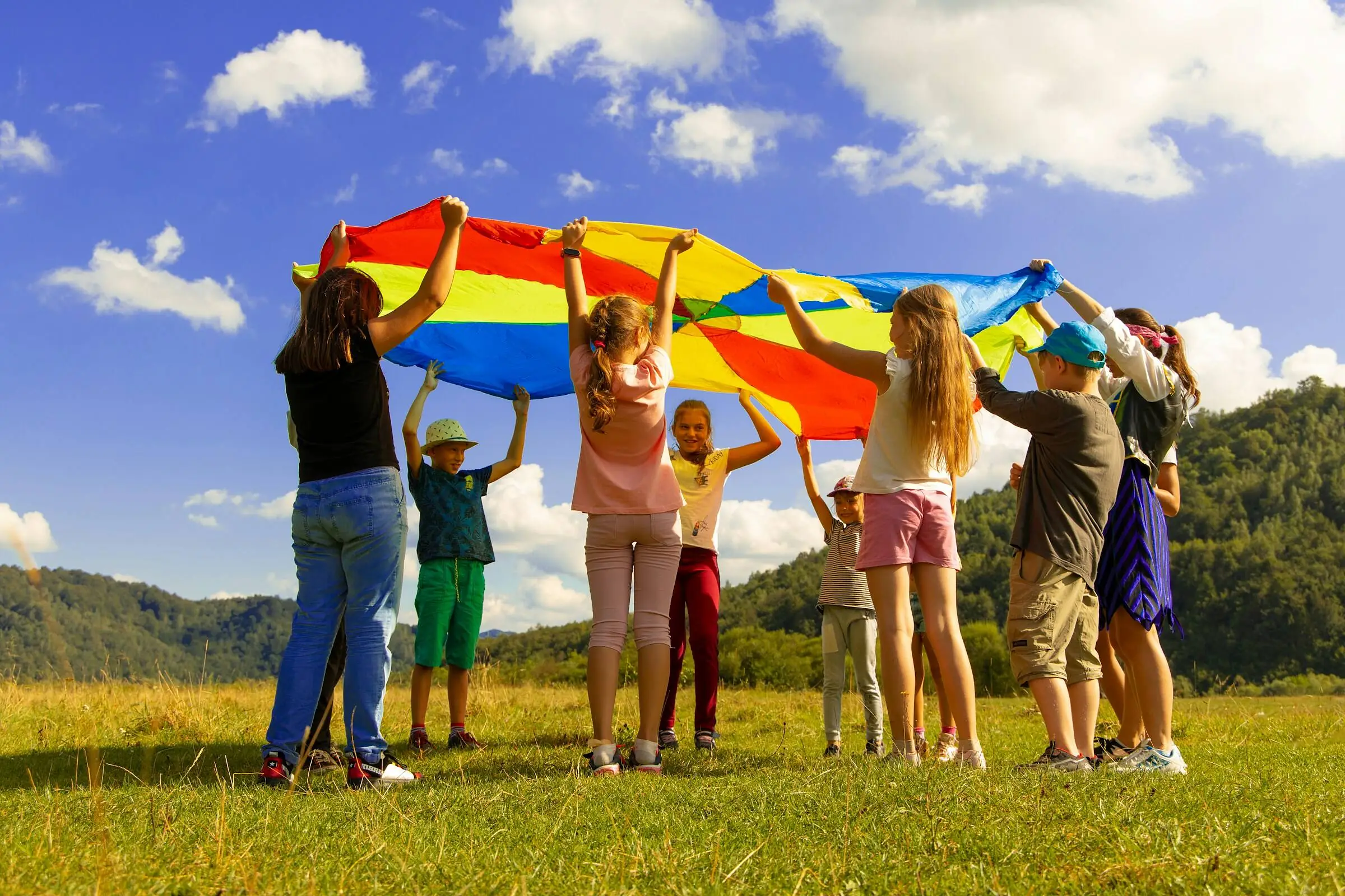 Children lifting a rainbow play parachute over their heads in a sunny meadow